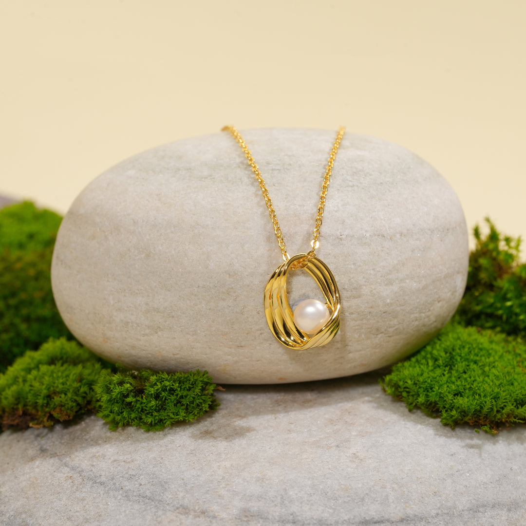 A contemporary gold necklace from Zewory Jewellery, featuring a sculptural cage pendant with a central pearl. The elegant piece is displayed on a smooth, light-grey stone and surrounded by fresh green moss for a natural aesthetic.
