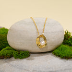 Load image into Gallery viewer, A contemporary gold necklace from Zewory Jewellery, featuring a sculptural cage pendant with a central pearl. The elegant piece is displayed on a smooth, light-grey stone and surrounded by fresh green moss for a natural aesthetic.
