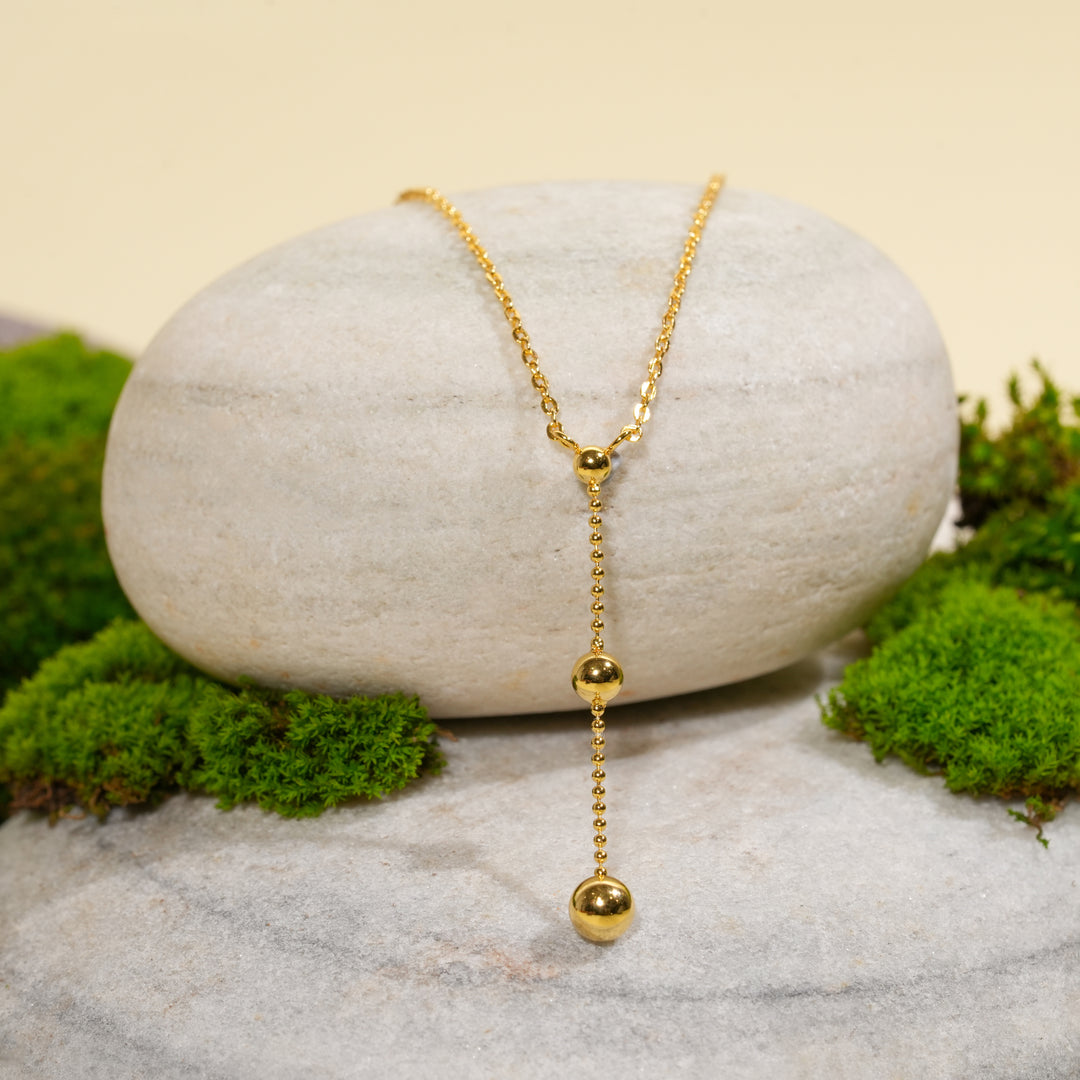 A modern gold lariat necklace from Zewory Jewellery, displayed in a serene natural setting. The elegant Y-shaped design, with its dangling beaded chain, is draped over a smooth, light-grey stone and surrounded by fresh green moss.