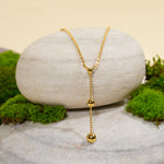 Load image into Gallery viewer, A modern gold lariat necklace from Zewory Jewellery, displayed in a serene natural setting. The elegant Y-shaped design, with its dangling beaded chain, is draped over a smooth, light-grey stone and surrounded by fresh green moss.
