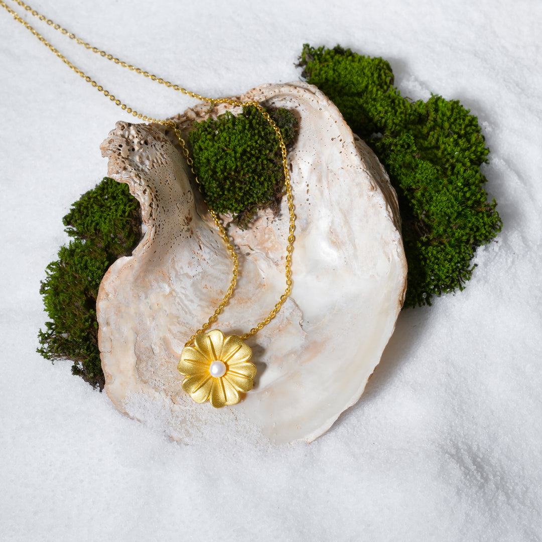 A stunning product shot of a gold flower and pearl necklace from Zewory Jewellery. This beautiful piece of botanical jewellery is artistically arranged inside a large seashell on a bed of white sand and moss, creating a luxurious image.