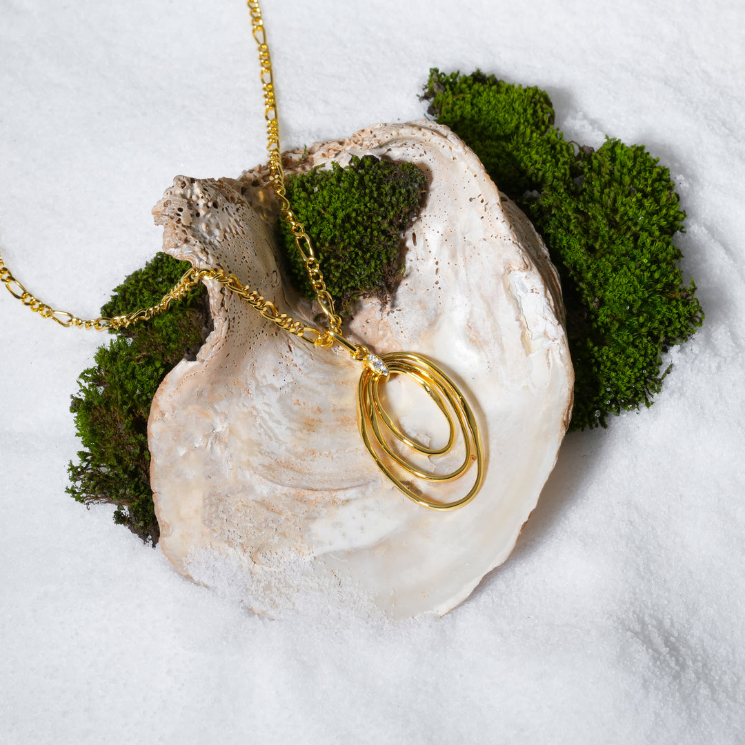 A luxurious product shot of a gold statement necklace from Zewory Jewellery. The pendant, featuring three interlocking oval hoops and a single crystal accent, is artfully displayed in a large seashell on a bed of white sand and moss.