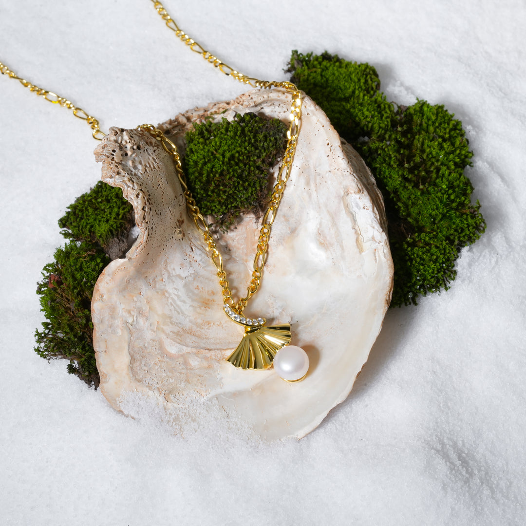  luxurious product shot of a gold ginkgo leaf and pearl necklace. This beautiful piece of botanical jewellery from Zewory is artistically arranged inside a large seashell on a bed of white sand and moss.