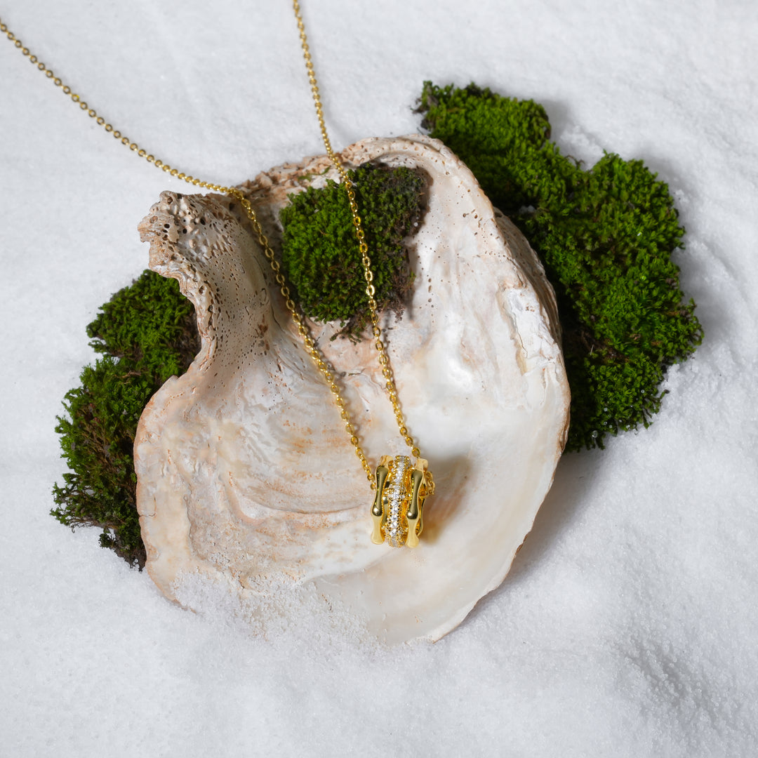 A luxurious product shot of a gold necklace featuring a barrel-shaped pendant with a pavé crystal center. This beautiful piece from Zewory Jewellery is artistically arranged inside a large seashell on a bed of white sand and moss.
