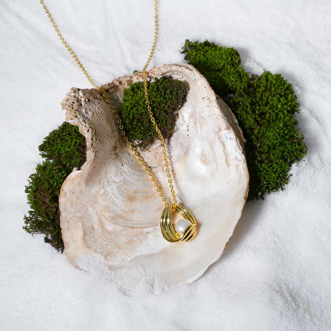 A luxurious product shot of a gold and pearl necklace featuring a sculptural cage pendant. This beautiful piece of contemporary jewellery from Zewory is artistically arranged inside a large seashell on a bed of white sand and moss.