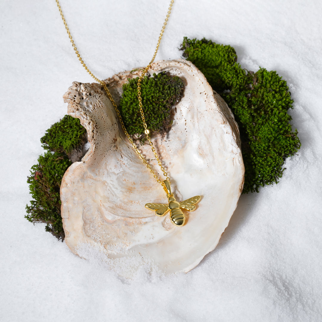 A stunning product shot of a gold bee pendant necklace. This beautiful piece of Zewory Jewellery is artistically arranged inside a large seashell on a bed of white sand and moss, creating a luxurious, nature-inspired image.