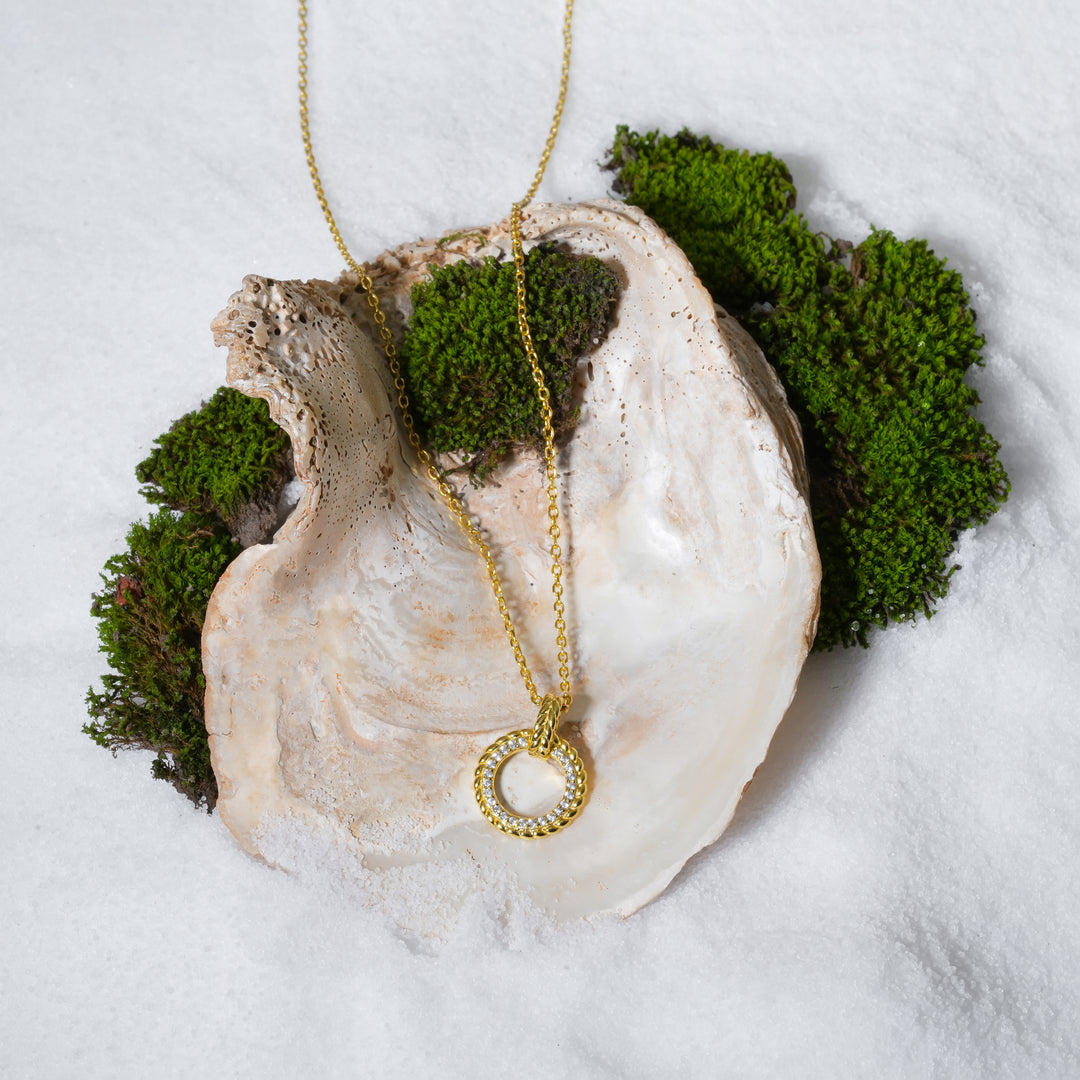 A luxurious product shot of a gold and crystal circle necklace from Zewory Jewellery. The elegant pendant, featuring a twisted rope texture and a pavé crystal half, is artistically arranged inside a large seashell on a bed of white sand and moss.