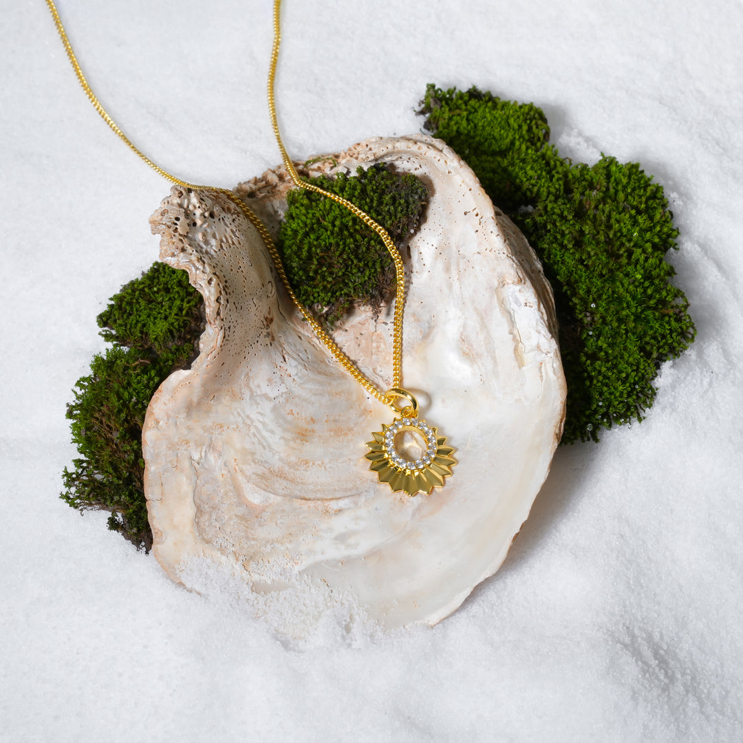 A luxurious product shot of a gold sun necklace with a crystal-lined center. This beautiful piece of celestial jewellery from Zewory is artistically arranged inside a large seashell on a bed of white sand and moss.