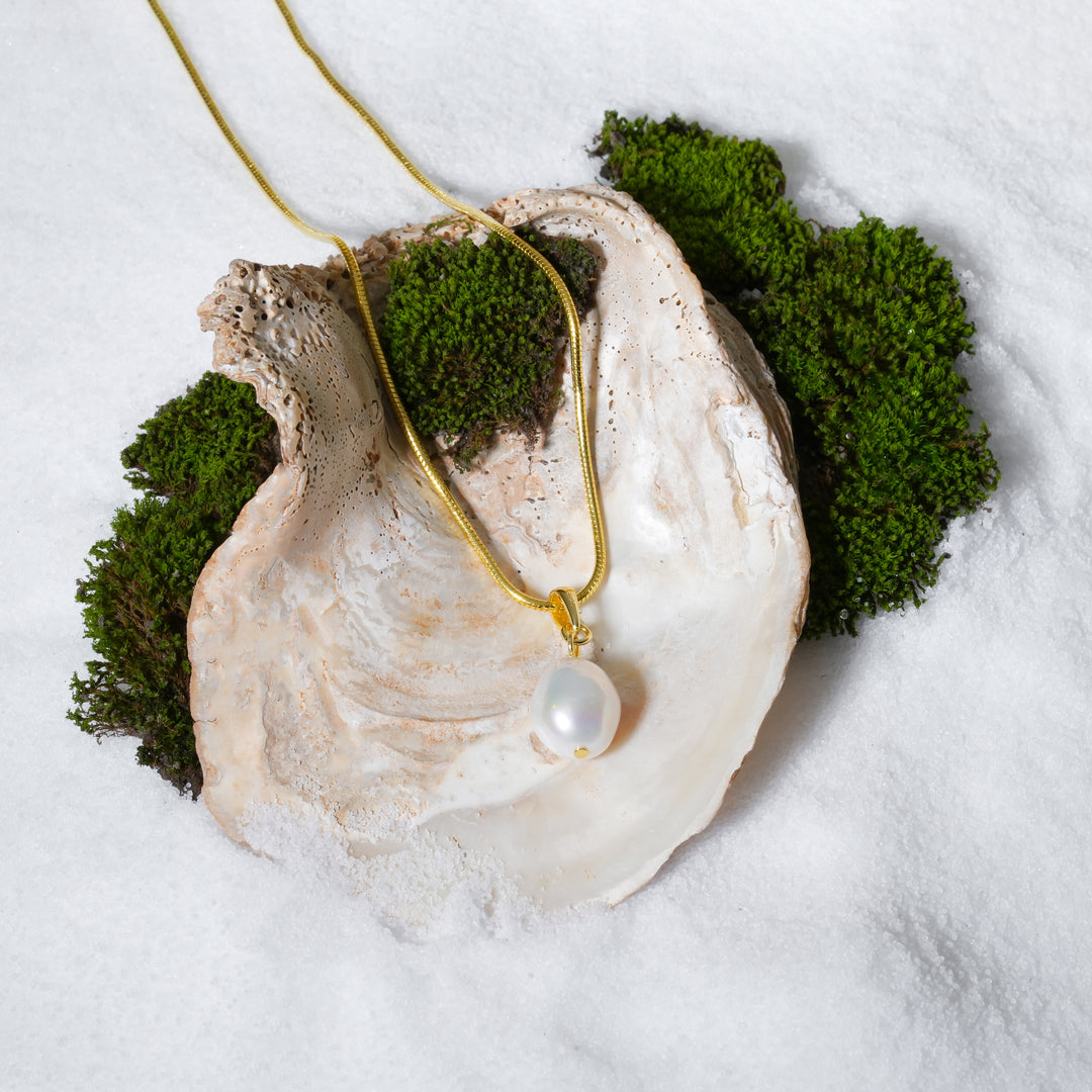A top-down view of a gold snake chain necklace with a single baroque pearl pendant. The necklace is artistically displayed inside a large seashell resting on a bed of white sand and accented with green moss.