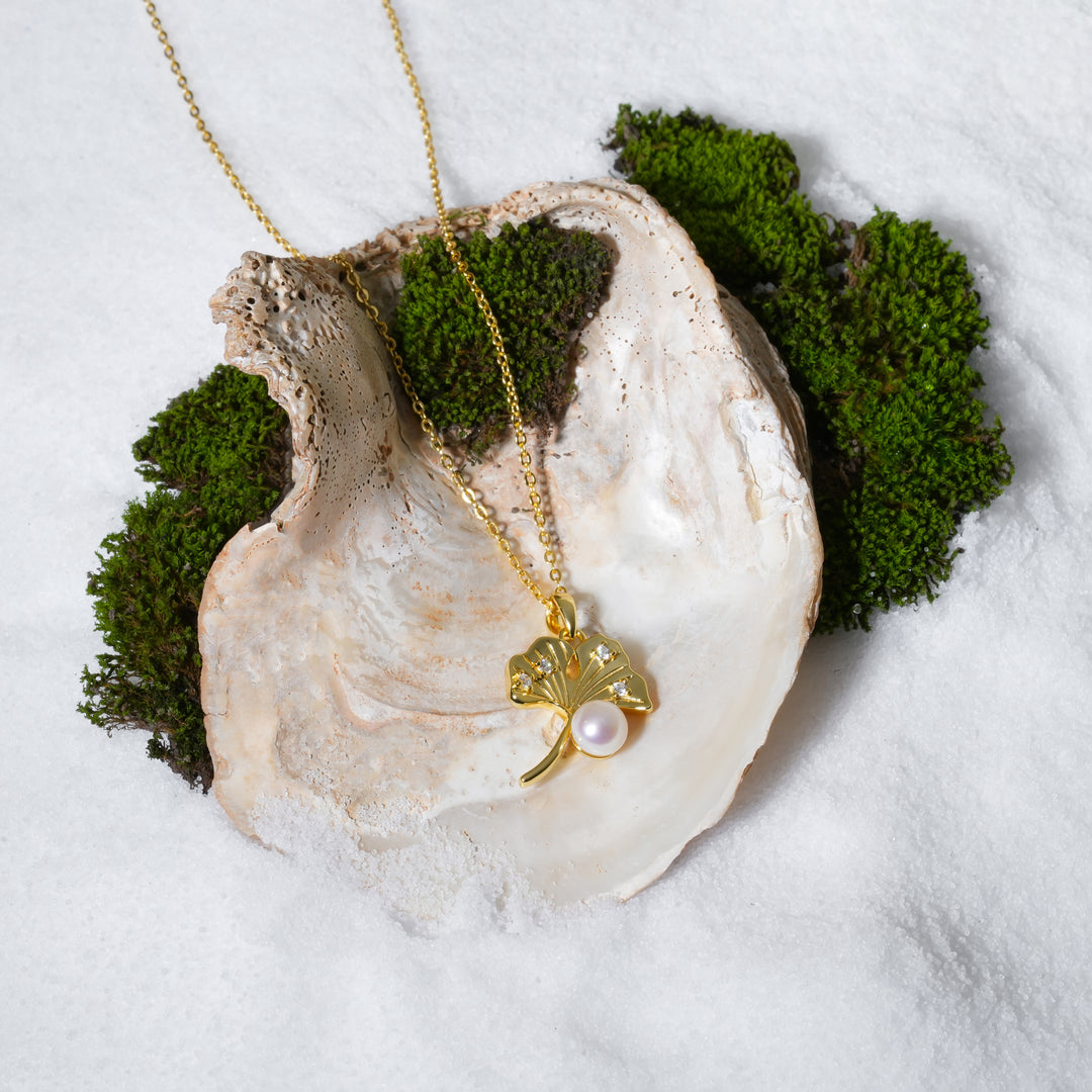 A gold necklace with a ginkgo leaf pendant is displayed inside a large, open seashell. The pendant features a white pearl and small crystal accents. The shell rests on a bed of white sand with vibrant green moss peeking from behind.