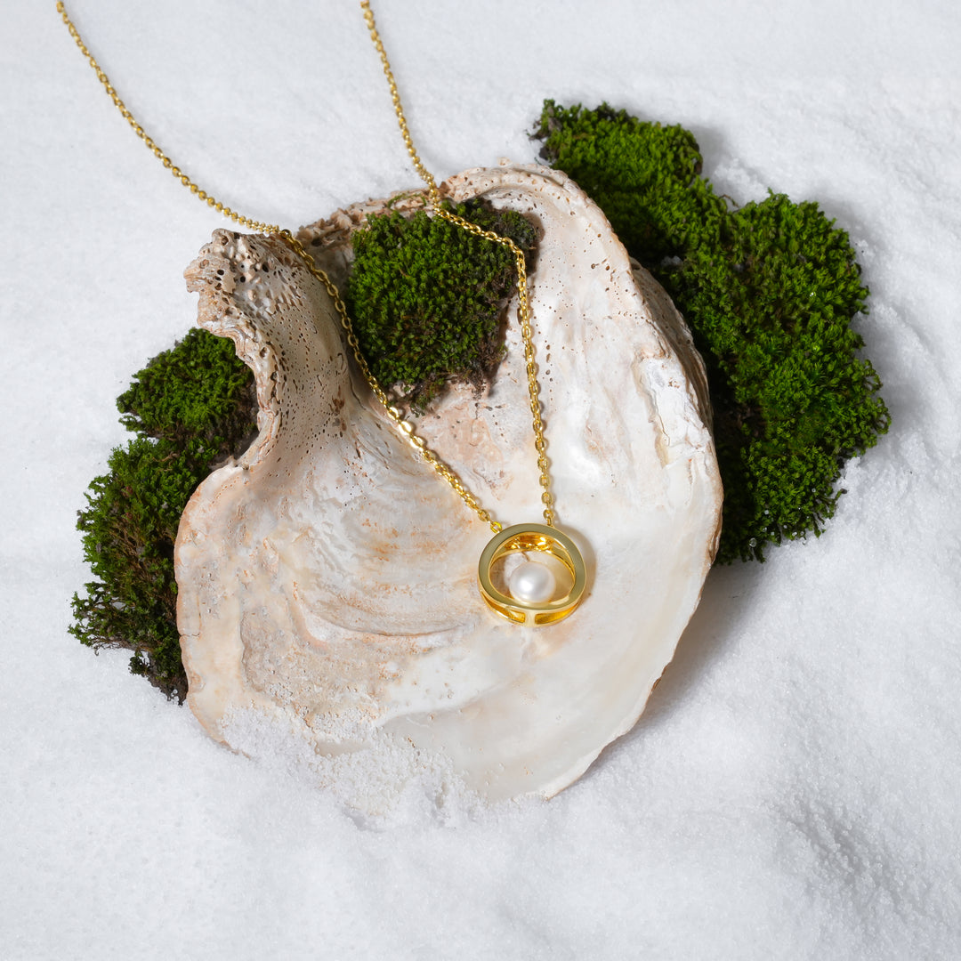 A luxurious product shot of a gold and pearl circle necklace. This beautiful piece of minimalist jewellery from Zewory is artistically arranged inside a large seashell on a bed of white sand, highlighting its modern elegance.