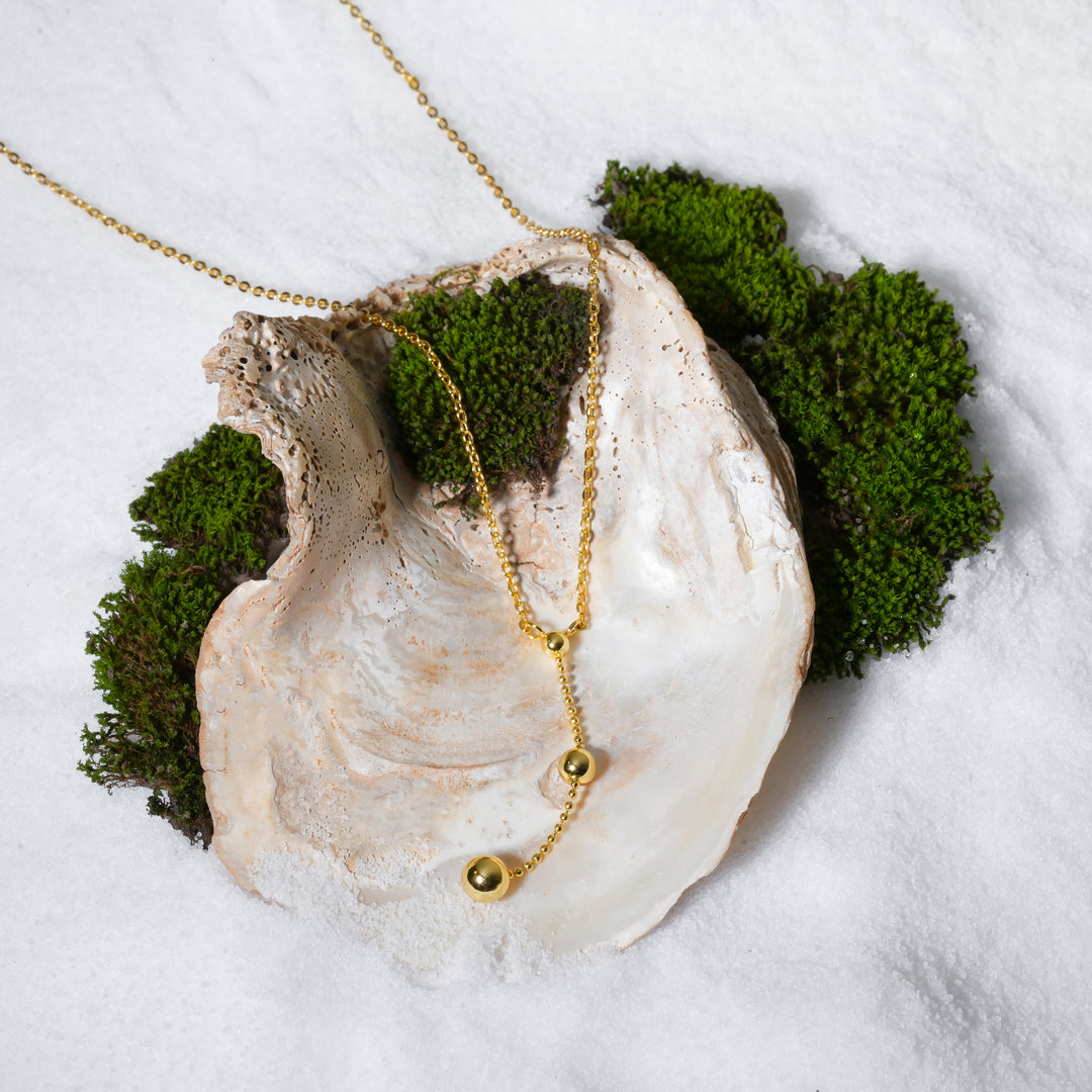 A luxurious product shot of a gold lariat-style necklace from Zewory Jewellery. This beautiful Y-necklace, featuring a drop chain with polished gold beads, is artistically arranged inside a large seashell on a bed of white sand and moss.