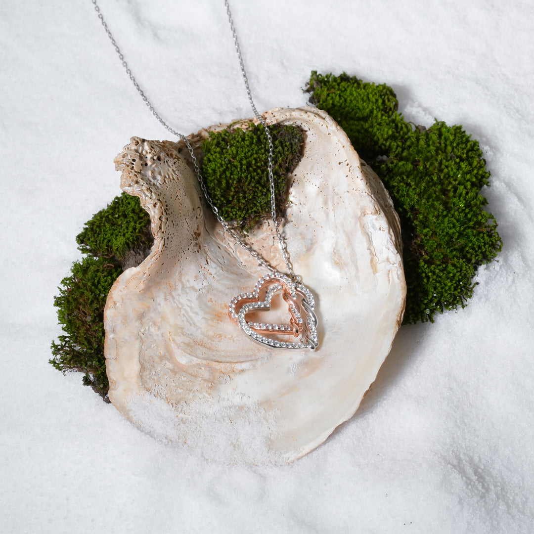 A luxurious product shot of a silver and rose gold interlocking hearts necklace. This beautiful and romantic piece from Zewory Jewellery is artistically arranged inside a large seashell on a bed of white sand, highlighting its sparkling crystal details.