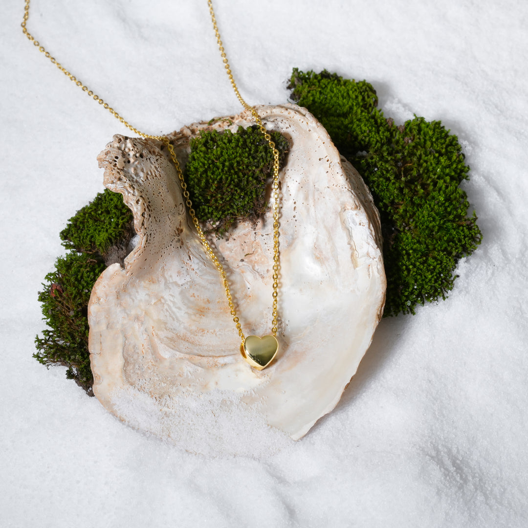 A luxurious product shot of a gold heart necklace from Zewory Jewellery. This beautiful and romantic piece, featuring a small polished heart, is artistically arranged inside a large seashell on a bed of white sand and moss.