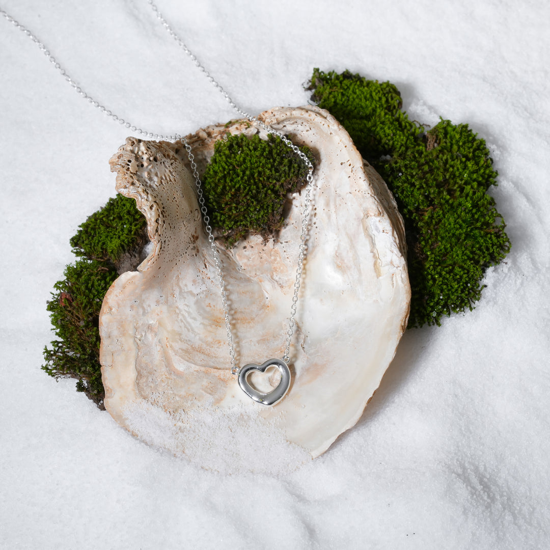 A luxurious product shot of a silver open heart necklace from Zewory Jewellery. This beautiful and romantic piece is artistically arranged inside a large seashell on a bed of white sand, highlighting its delicate charm.