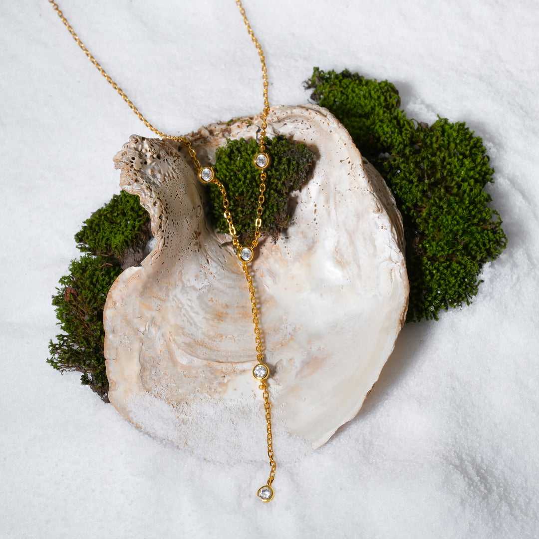 A luxurious product shot of a gold lariat necklace from Zewory Jewellery. This delicate Y-necklace, sparkling with bezel-set crystals, is artistically arranged inside a large seashell on a bed of white sand and moss.