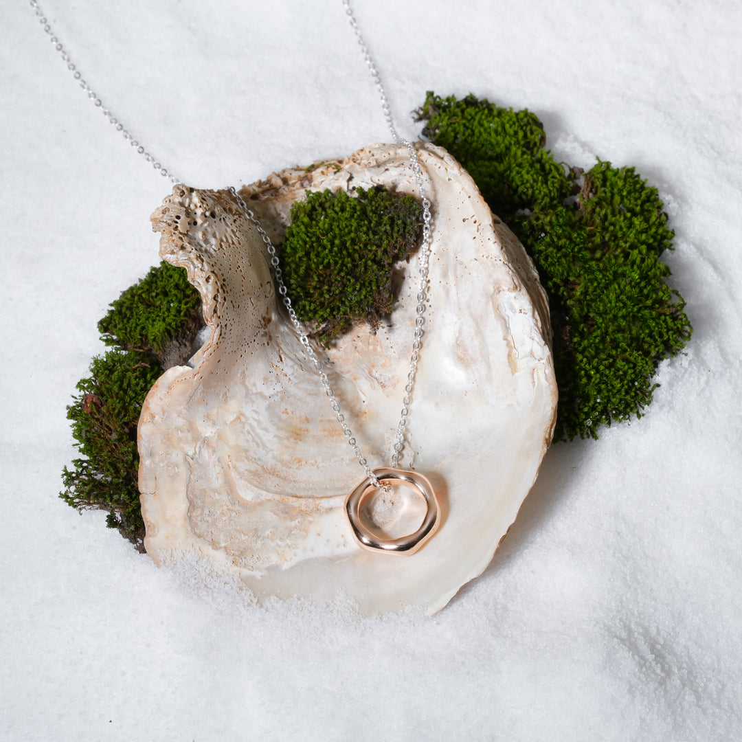 A luxurious product shot of a silver and rose gold necklace. This beautiful piece of mixed-metal jewellery from Zewory, with its organic circle pendant, is artistically arranged inside a large seashell on a bed of white sand and moss.