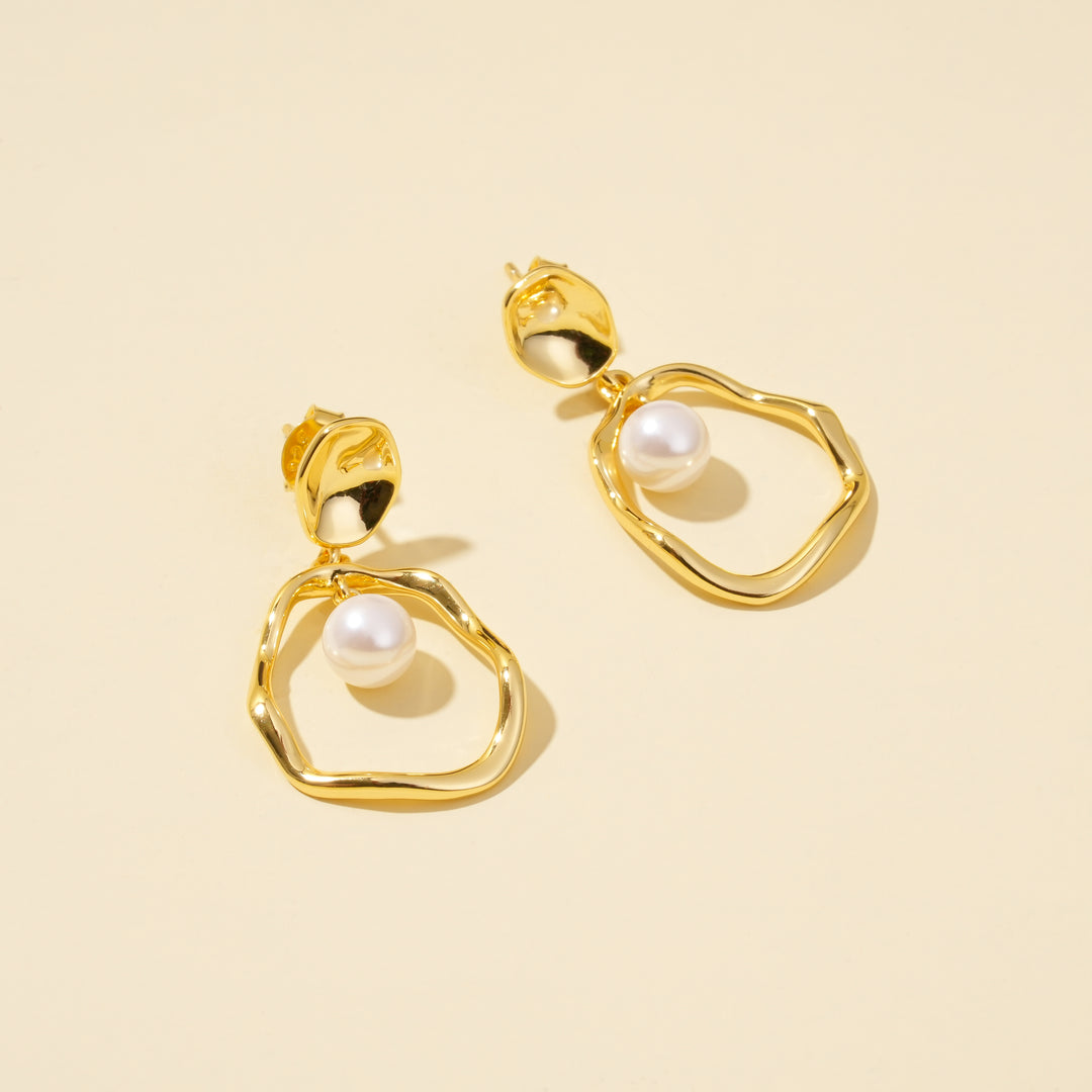 A top-down product shot of elegant gold and pearl drop earrings from Zewory Jewellery. The image clearly shows the unique design, with a single pearl dangling inside each organic-shaped gold hoop, on a clean, neutral background.