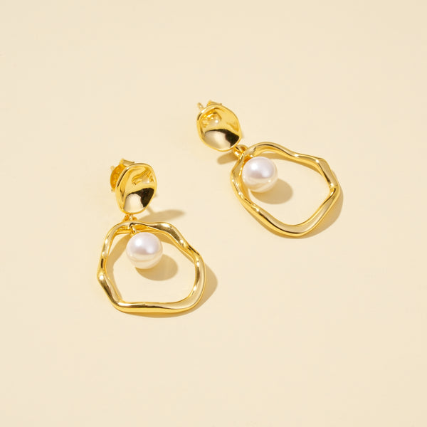 A top-down product shot of elegant gold and pearl drop earrings from Zewory Jewellery. The image clearly shows the unique design, with a single pearl dangling inside each organic-shaped gold hoop, on a clean, neutral background.