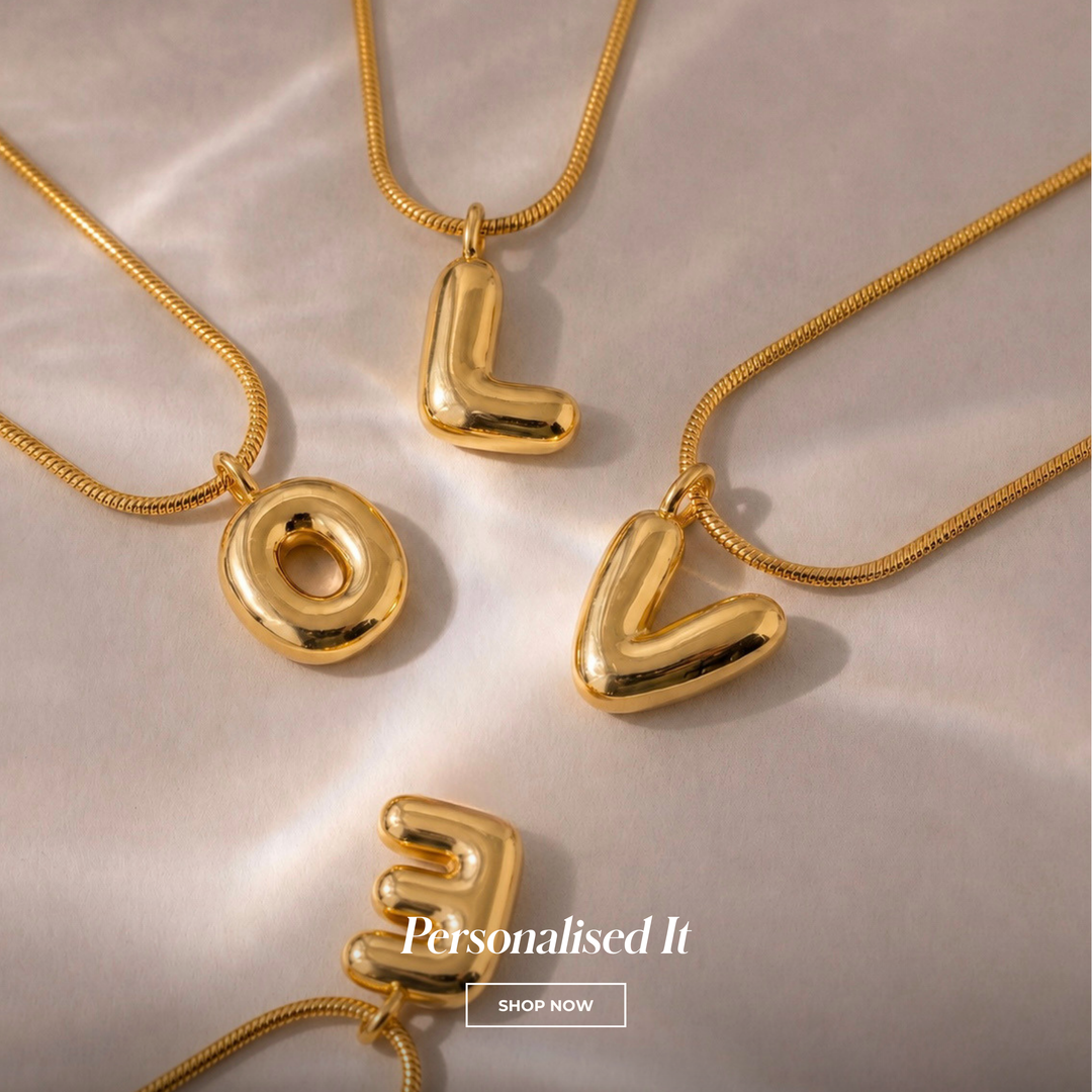 Four gold letter necklaces spelling LOVE are arranged on a light beige satin fabric background. The letters are polished and metallic, suspended from thin snake-chain necklaces.