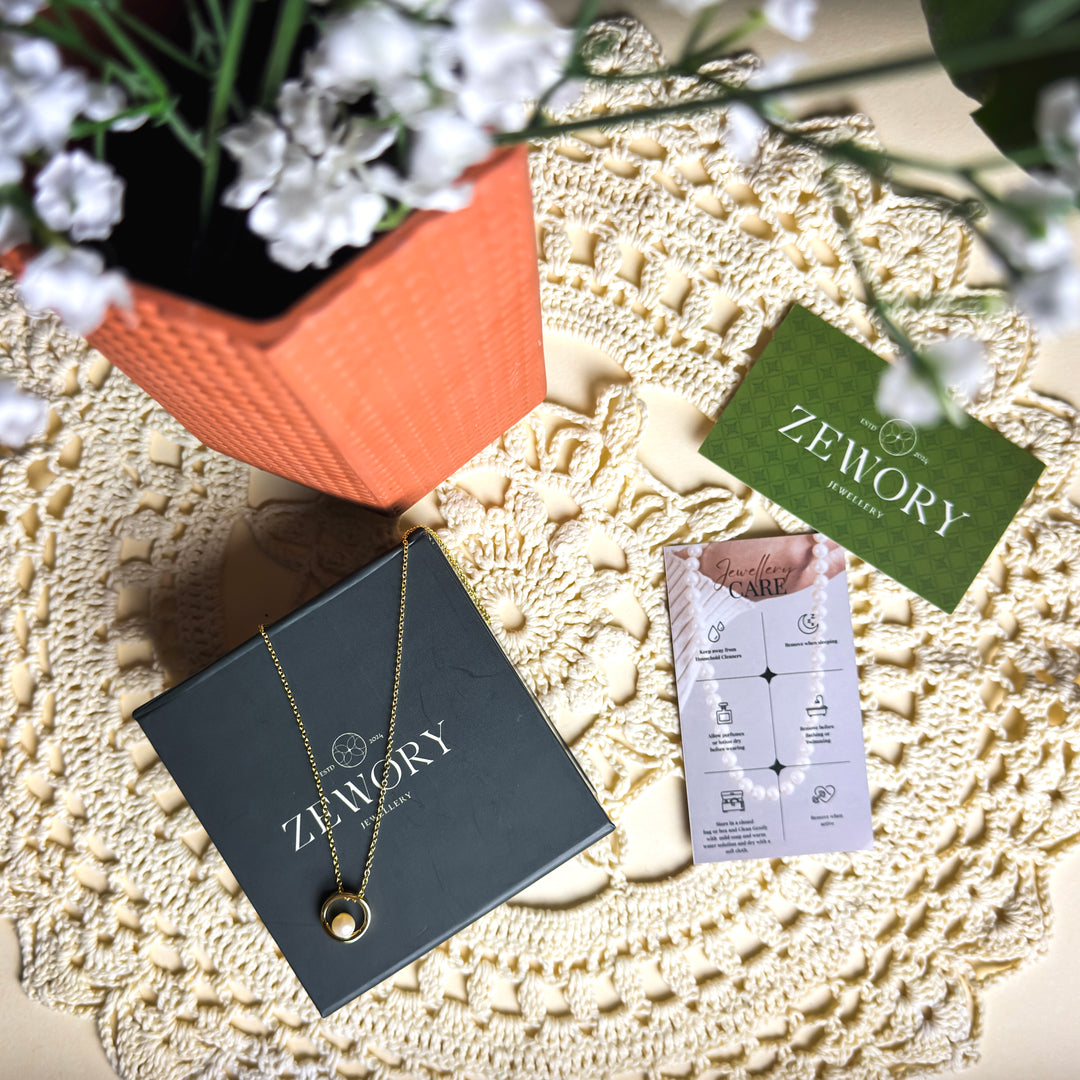 Zewory necklace on a dark green box with a card and tag on a textured surface with flowers.