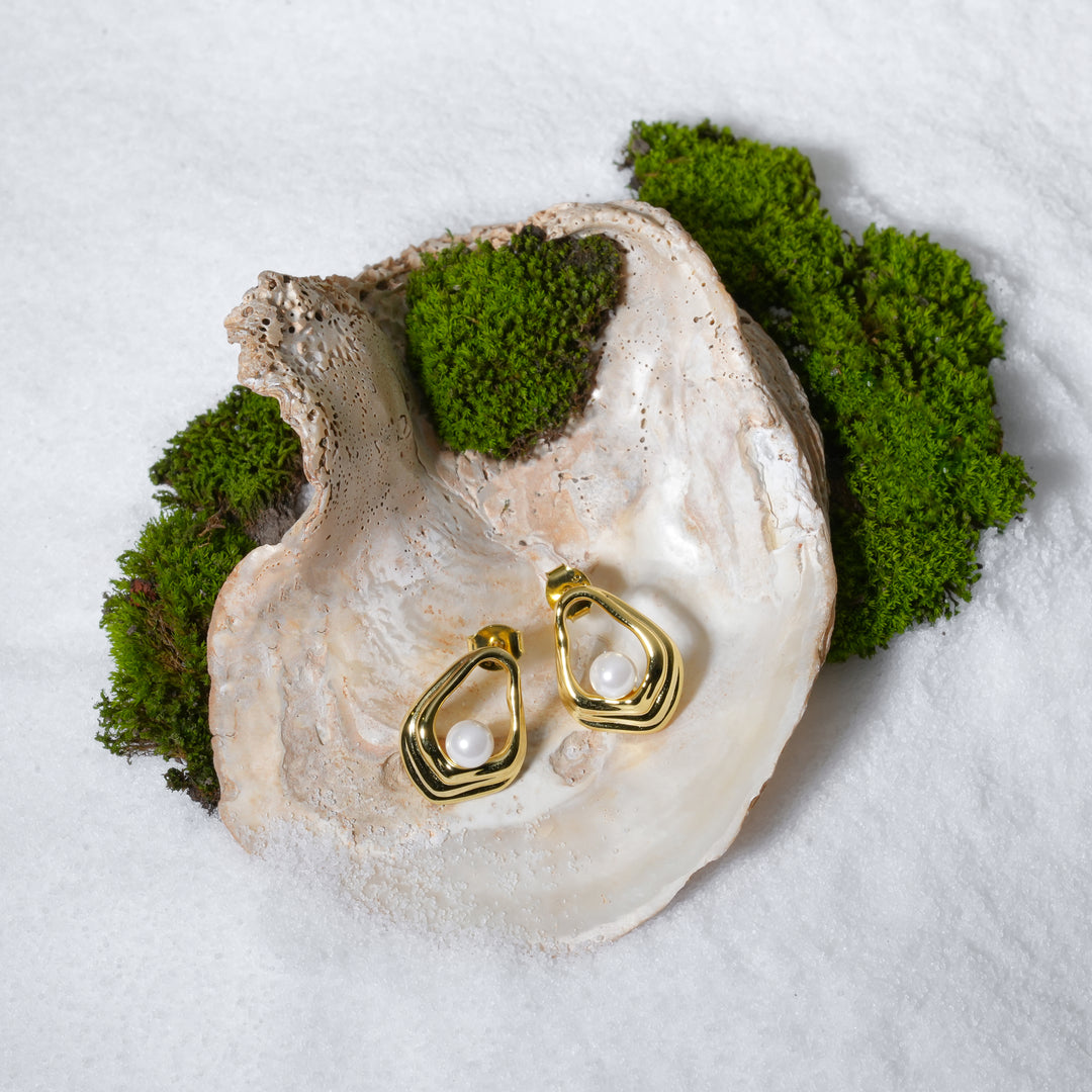 An artistic product shot of modern gold and pearl stud earrings from Zewory Jewellery. The pair of abstract, open-design studs are elegantly displayed inside a large seashell on a bed of white sand and moss.