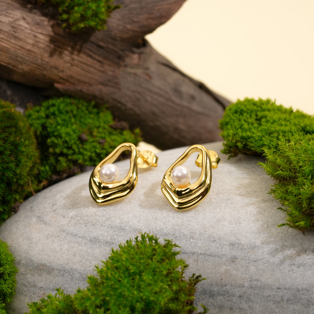 A pair of contemporary gold and pearl stud earrings displayed in a natural setting. The organic, open-frame design of each earring holds a single pearl, beautifully showcased on a grey stone with moss and weathered wood.