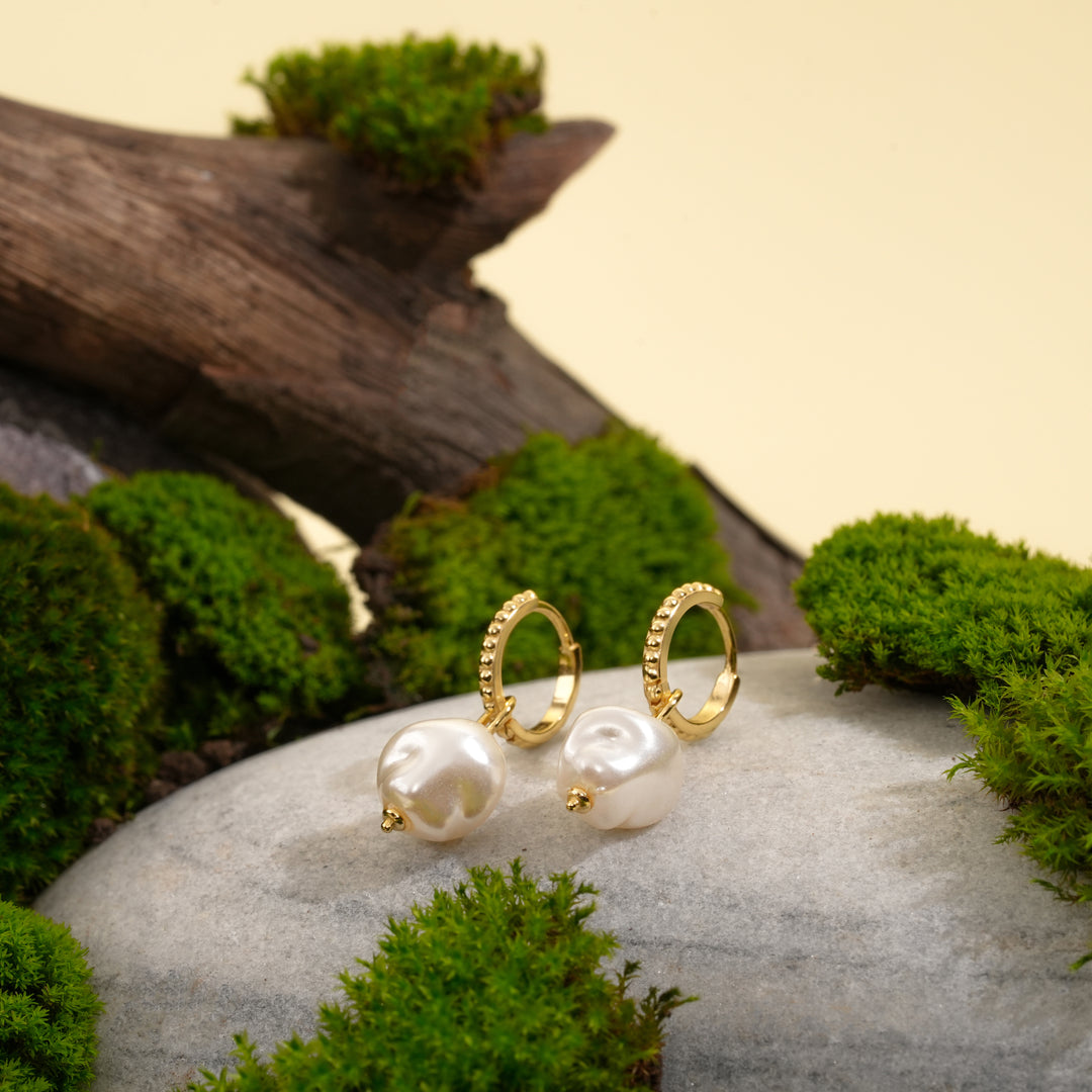A pair of elegant gold baroque pearl huggie earrings from Zewory Jewellery, displayed in a natural setting on a grey stone with green moss and wood.