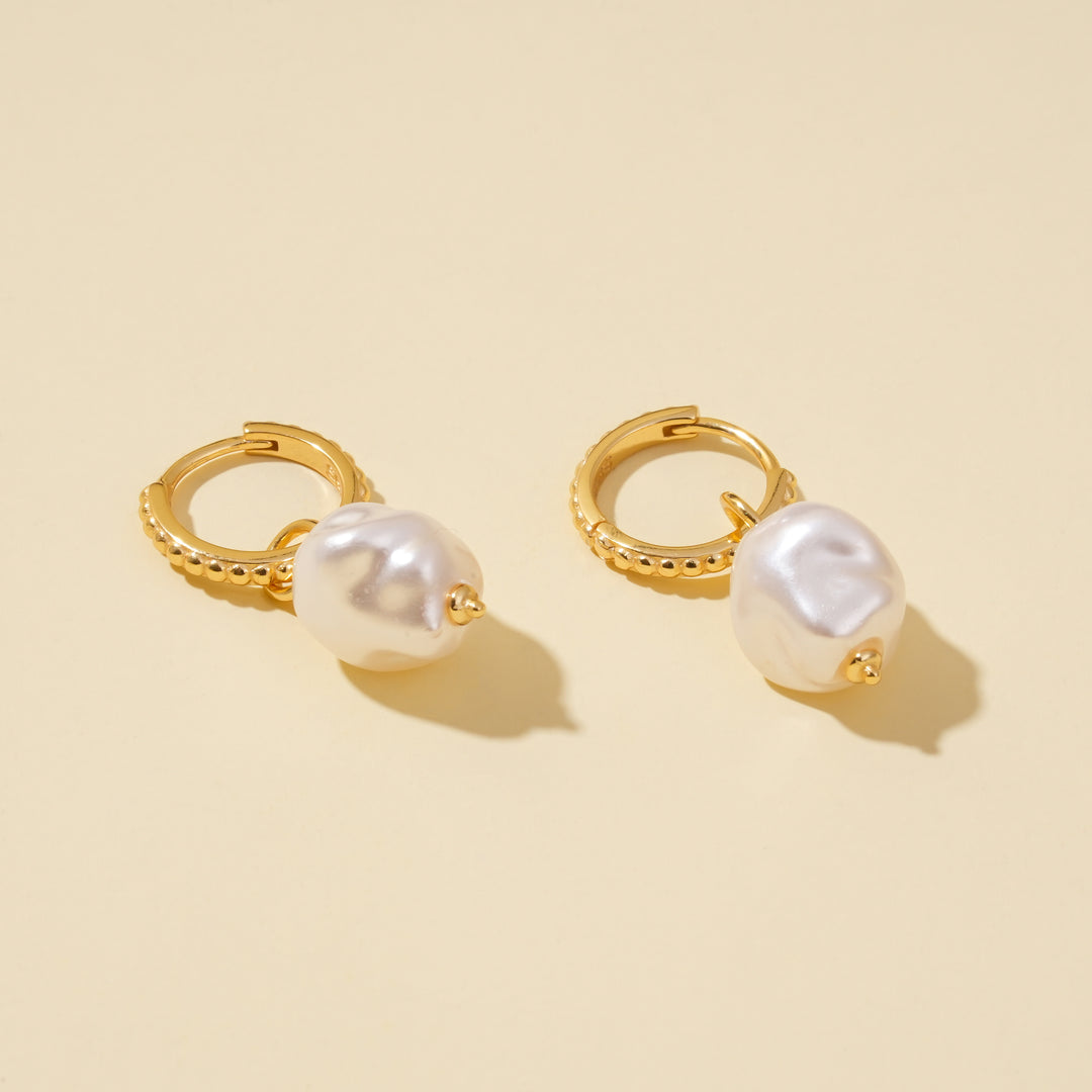 A clean product shot of a pair of gold huggie hoop earrings with large, dangling baroque pearls, set against a neutral cream background.