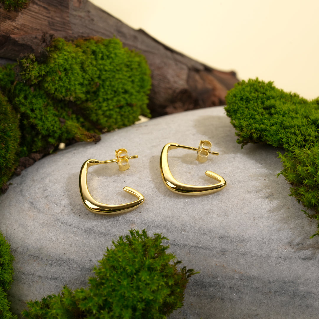 A pair of minimalist, C-shaped gold hoop earrings from Zewory Jewellery are displayed on a smooth grey stone. The modern, geometric design is highlighted by a natural setting of vibrant green moss and dark wood.