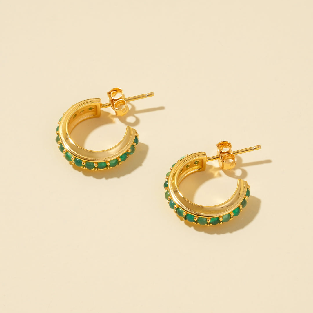 A clean product shot of a pair of gold C-hoop earrings, showcasing the channel-set, vibrant green gemstones on a neutral cream background.