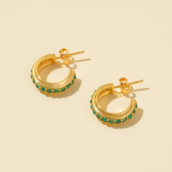 A clean product shot of a pair of gold C-hoop earrings, showcasing the channel-set, vibrant green gemstones on a neutral cream background.