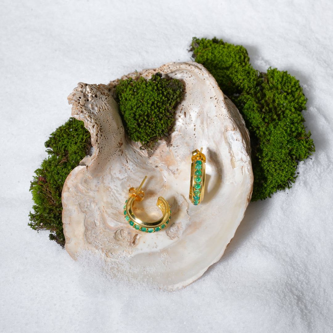 A luxurious product shot of gold and emerald-green hoop earrings, artistically displayed inside a large seashell on a bed of white sand and moss.