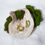Load image into Gallery viewer, A luxurious product shot of gold and emerald-green hoop earrings, artistically displayed inside a large seashell on a bed of white sand and moss.
