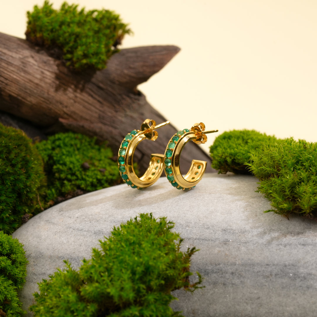 A pair of gold and green gemstone hoop earrings from Zewory Jewellery, displayed in a natural setting on a grey stone with green moss and weathered wood.