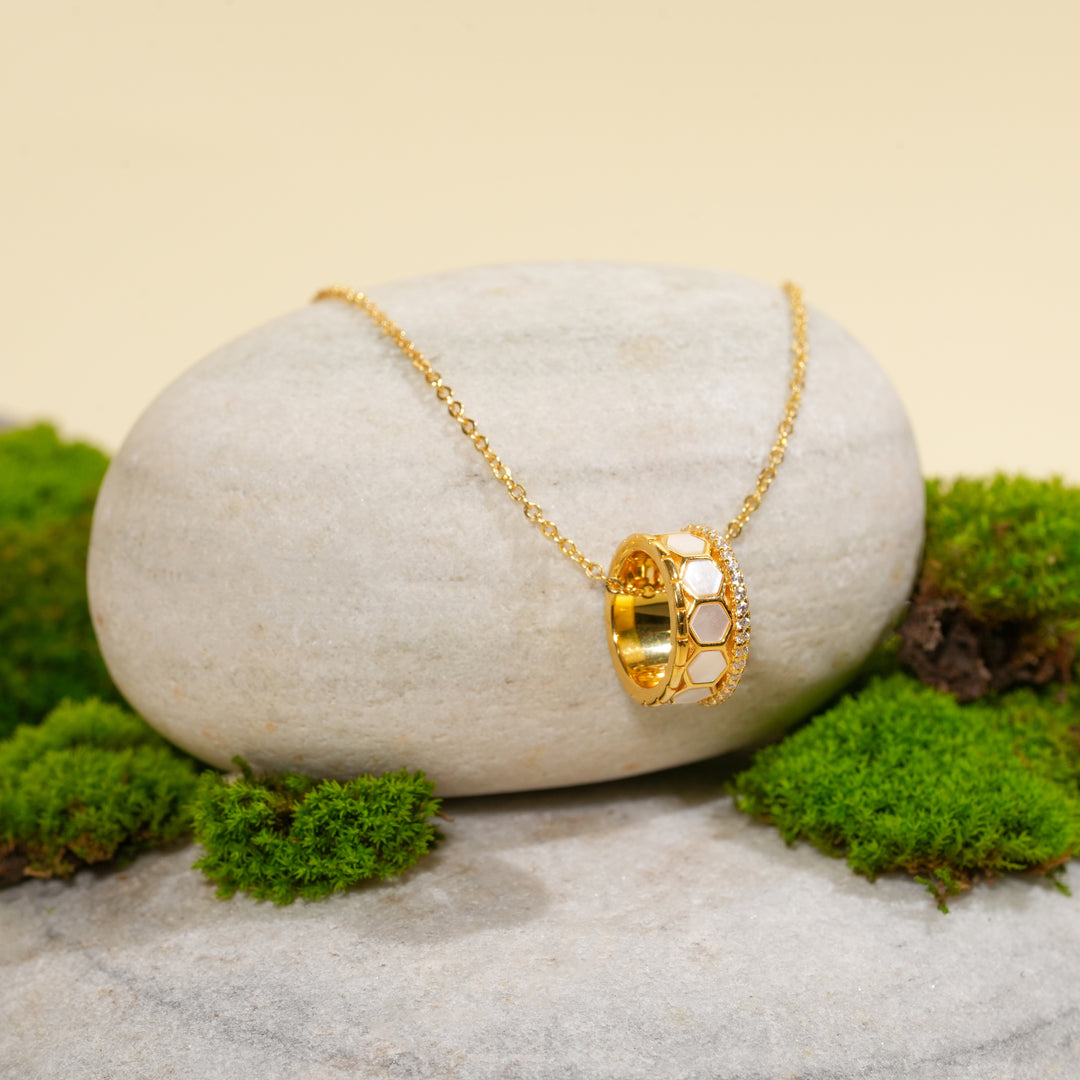 A modern gold honeycomb necklace from Zewory Jewellery, displayed in a serene natural setting. The unique barrel pendant, featuring mother-of-pearl inlays and a crystal accent band, is draped over a smooth, light-grey stone and surrounded by fresh green moss.