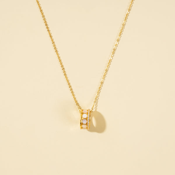 A clean, minimalist product shot of a gold honeycomb necklace on a neutral cream background. The image clearly displays the intricate barrel pendant, which features white mother-of-pearl hexagonal inlays bordered by a band of sparkling pavé crystals.