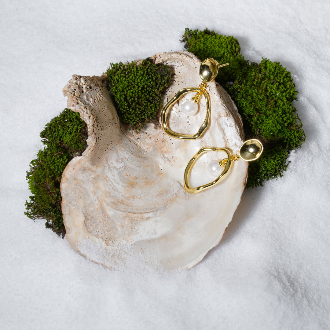 A pair of luxurious gold and pearl dangle earrings from Zewory Jewellery, elegantly displayed on a large seashell. The modern, organic-shaped hoops and lustrous pearls are set against a backdrop of white sand and moss for a beautiful product shot.