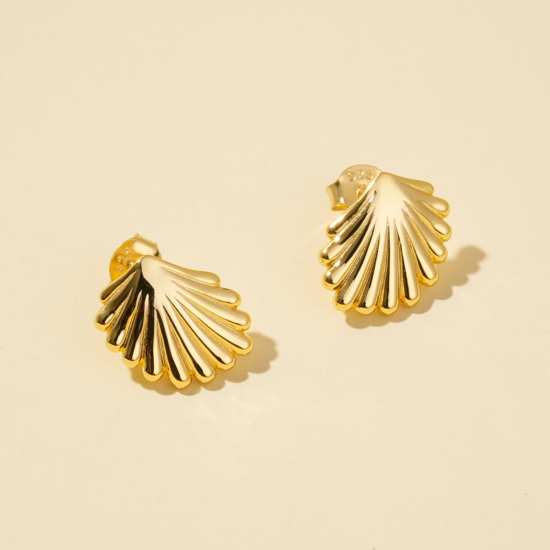 A clean, minimalist product shot of a pair of gold seashell stud earrings on a neutral cream background. The image clearly displays the intricate, ribbed texture of the classic scallop shell design from an overhead angle.