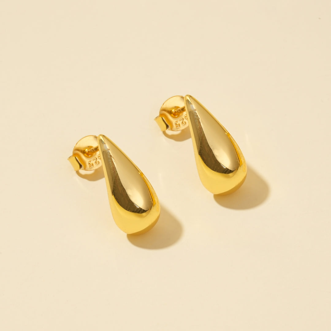 A clean product shot of a pair of modern gold teardrop stud earrings. The minimalist, polished droplet design is clearly displayed against a neutral cream background, showing their elegant and simple form from an overhead angle.