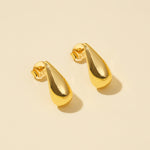 Load image into Gallery viewer, A clean product shot of a pair of modern gold teardrop stud earrings. The minimalist, polished droplet design is clearly displayed against a neutral cream background, showing their elegant and simple form from an overhead angle.

