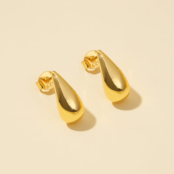 A clean product shot of a pair of modern gold teardrop stud earrings. The minimalist, polished droplet design is clearly displayed against a neutral cream background, showing their elegant and simple form from an overhead angle.