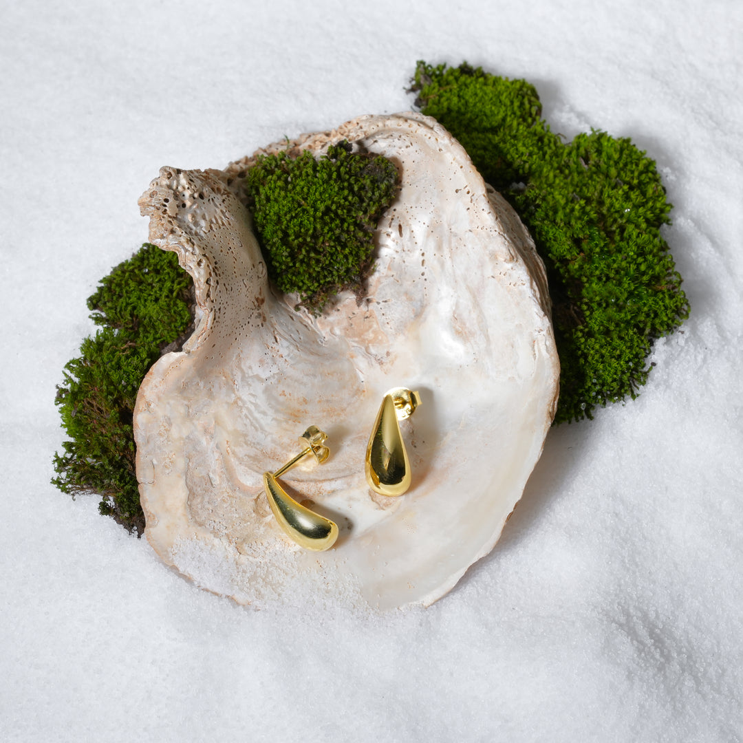 A luxurious product shot of minimalist gold teardrop stud earrings from Zewory Jewellery. The pair of elegant, polished droplet studs are artistically displayed inside a large seashell on a bed of white sand and moss.