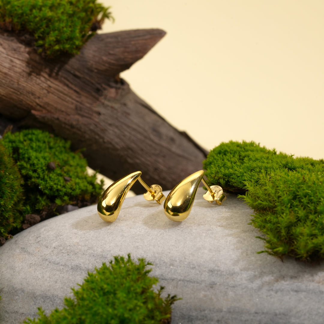 A pair of minimalist gold teardrop stud earrings from Zewory Jewellery. The modern, polished droplet design is showcased in a natural setting on a grey stone, surrounded by vibrant green moss and weathered wood.