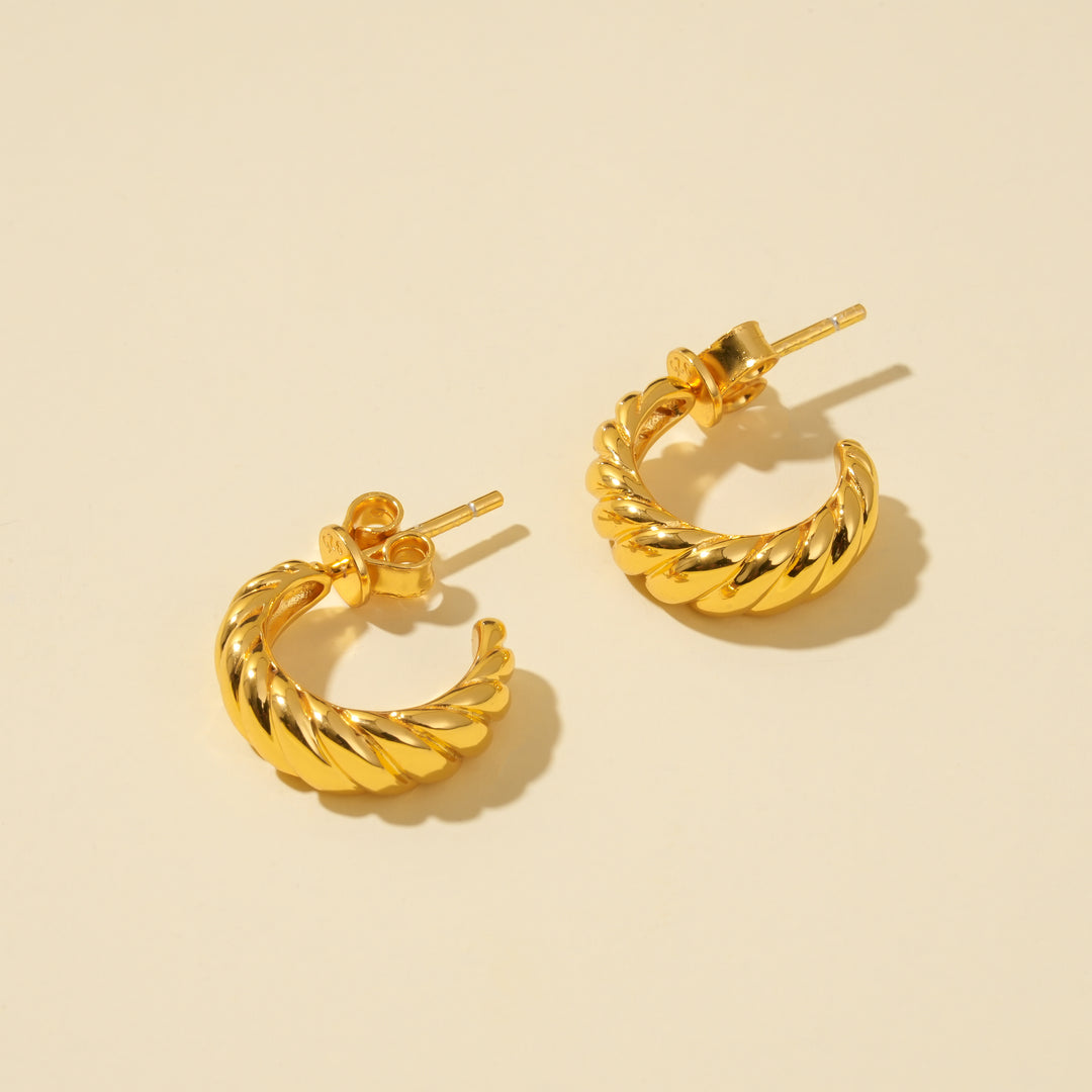 A clean, minimalist product shot of a pair of gold twisted hoop earrings on a neutral cream background. The image clearly displays the trendy, croissant-like texture and C-shape of the stylish hoops from an overhead angle.