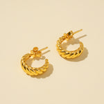 Load image into Gallery viewer, A clean, minimalist product shot of a pair of gold twisted hoop earrings on a neutral cream background. The image clearly displays the trendy, croissant-like texture and C-shape of the stylish hoops from an overhead angle.
