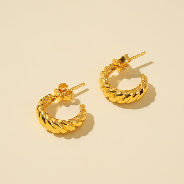 A clean, minimalist product shot of a pair of gold twisted hoop earrings on a neutral cream background. The image clearly displays the trendy, croissant-like texture and C-shape of the stylish hoops from an overhead angle.