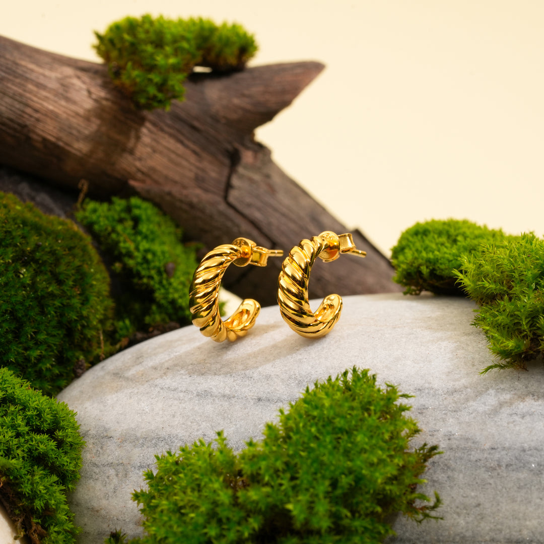 A pair of modern, twisted gold hoop earrings from Zewory Jewellery. The unique, rope-like texture is showcased in a natural setting on a grey stone, surrounded by vibrant green moss and weathered wood.
