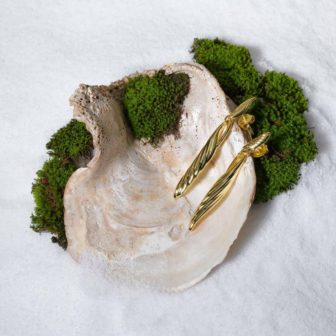 A pair of striking, twisted gold drop statement earrings from Zewory Jewellery, creatively displayed on a large seashell. The product is set on a bed of white sand and accented with vibrant green moss for a natural, luxurious aesthetic.