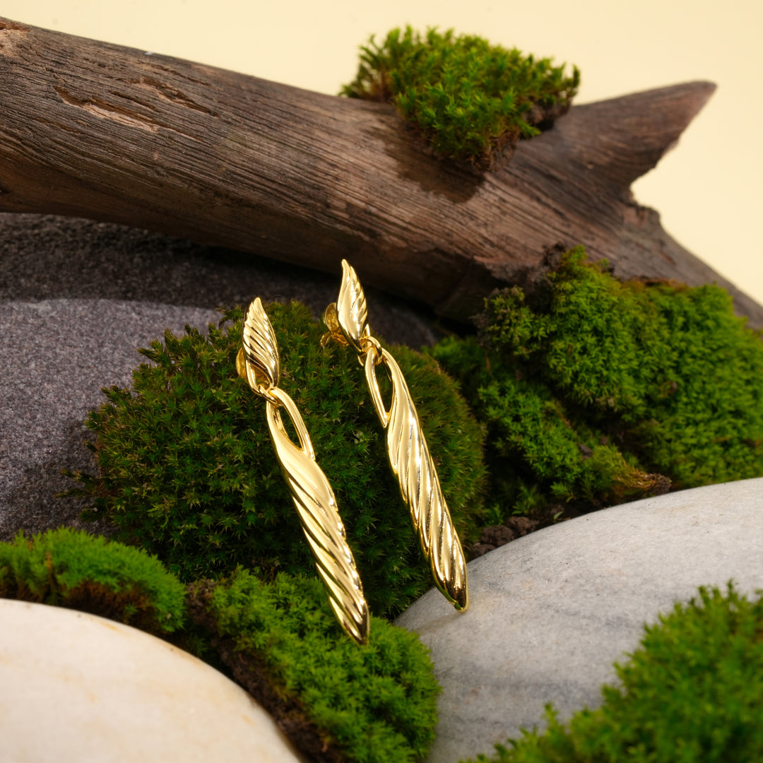 A pair of elegant twisted gold drop earrings showcased in an earthy setting. The earrings are propped against green moss, weathered wood, and smooth stones, highlighting their intricate design and polished finish.
